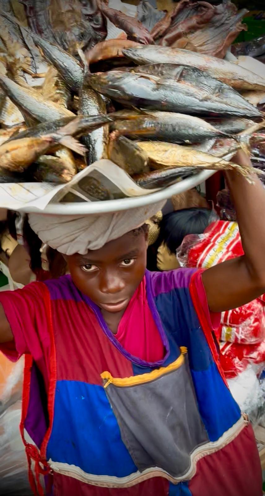 A young vendor carrying a tray of smoked fish through an Accra market